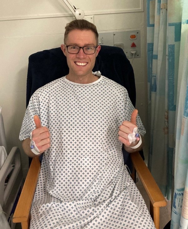 Living kidney donor Rich sitting in a chair wearing a hospital gown. He is grinning widely at the camera and giving two thumbs up.