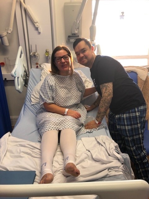 Living kidney donor Ashleigh with her donor husband and recipient in hospital. Ashleigh is in a hospital gown lying on a bed, leaning forward and smiling for the camera, while her husband is standing alongside her in comfy clothes, leaning in for the photo.