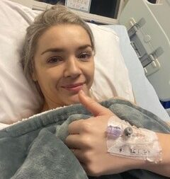 Living kidney donor Ellie lying in a hospital bed, with a blanket pulled up to her neck. She is giving a big smile and thumbs up for the camera