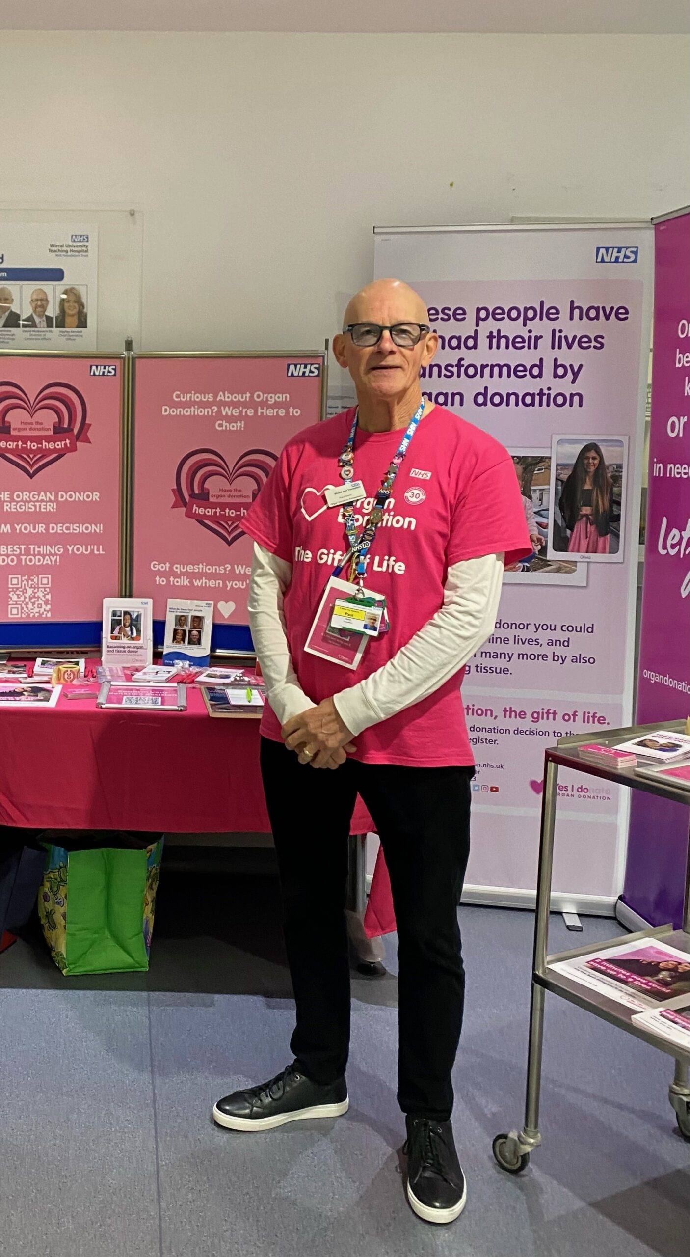 Living kidney donor Paul standing by an NHS Blood & Transplant stand. He is wearing a pink volunteer's top featuring the words 'Organ Donation: the Gift of Life' beside a picture of a heart.