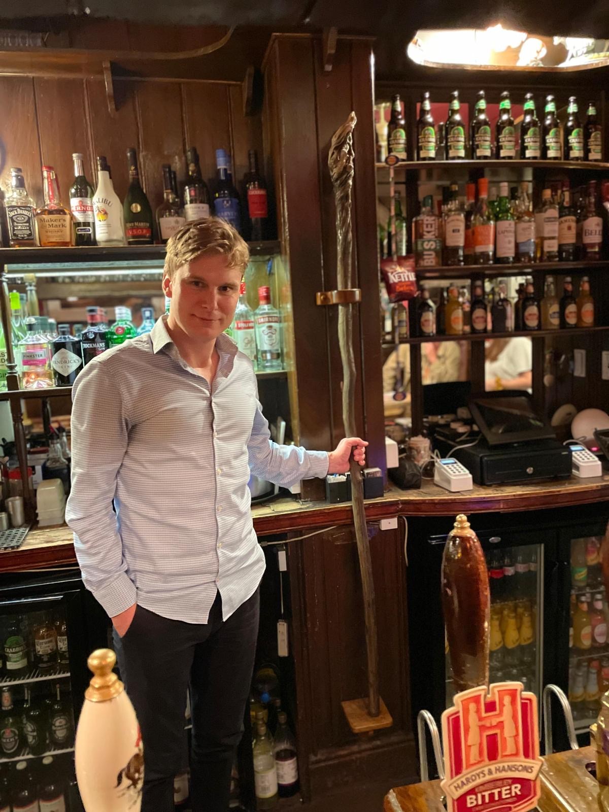 Image of living kidney donor Keegan standing by the bar in an old British pub. Beside him is the staff of Gandalf the Grey from the Lord of the Rings movie, propped up in a holder by the bar.