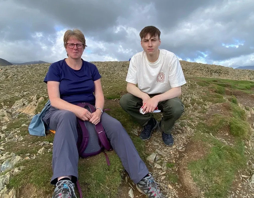 Living kidney recipient Dan alongside his mum Alison. They are crouching on a rocky hillside and both looking at the camera, rolling light grey clouds coming in behind them.