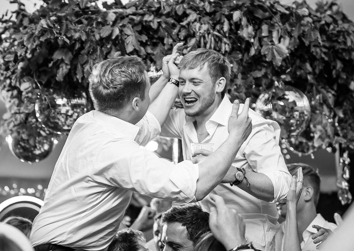 Black and white image of Ed and George at a wedding, both wearing a white shirts, leaning towards each other and laughing