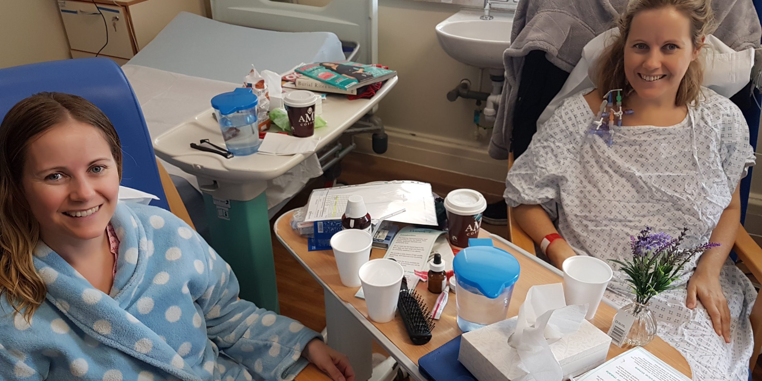 Image of living kidney donor Jennie with her sister and kidney recipient Sue. The two are in hospital, both wearing hospital gowns, with one of them in bed and the other sitting in a chair beside them, both smiling for the camera.