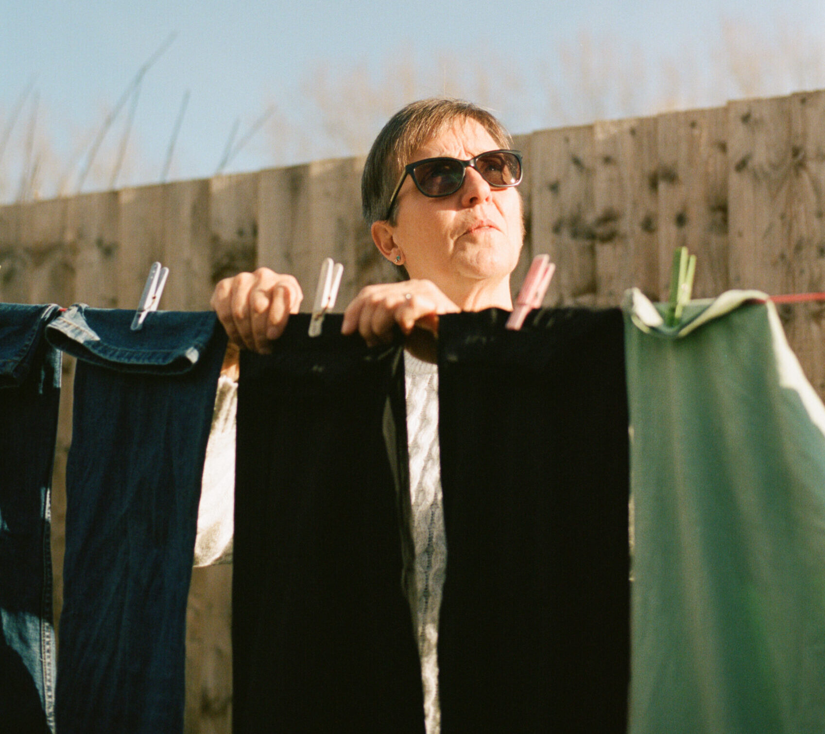 Living kidney donor Charlotte hanging out her washing on a washing line beneath a clear blue sky. She is wearing sunglasses and looking up at the horizon to her left.