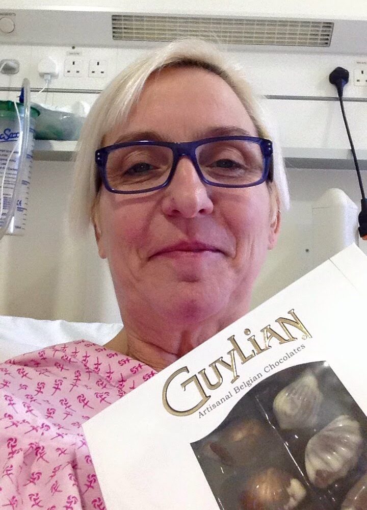 Image of living kidney donor Charlotte. She is in a hospital bed, with a row of plugs and sockets visible behind her. She is wearing black glasses and holding a box of Guylian chocolates in front of her, smiling.