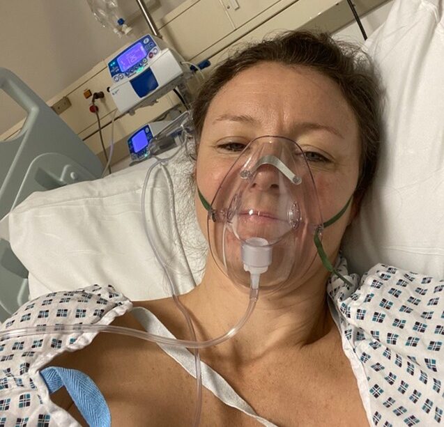 Selfie of living kidney donor Clare in a hospital bed, wearing a hospital gown and a transparent gas mask over her mouth.