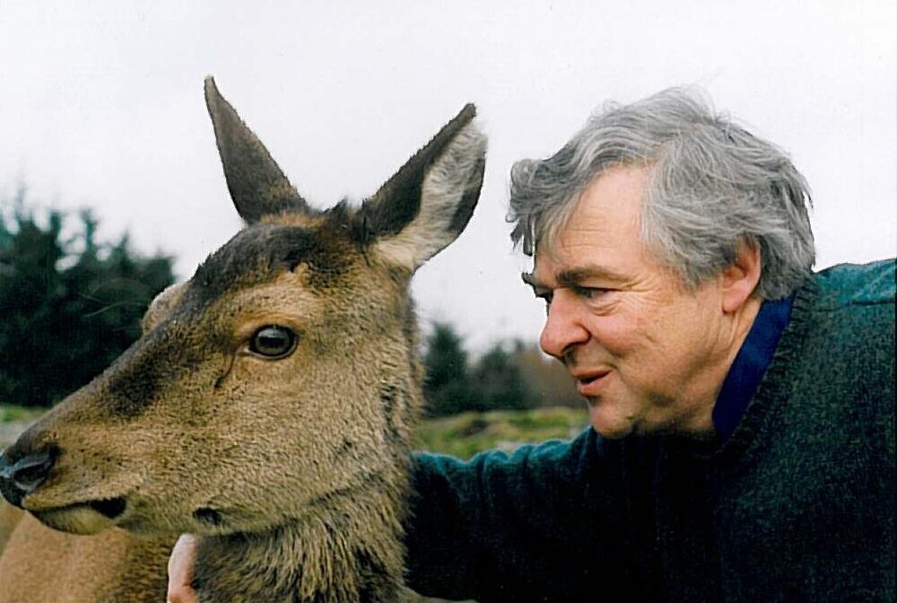 Image of living kidney donor John standing in a field with a deer close beside him. He is looking at the deer with a smile, while the animal is turned away looking at something to the left of the camera,