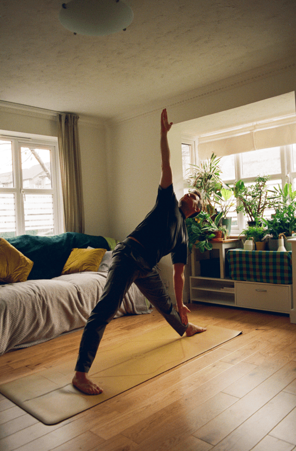 Living kidney donor Jenson. He is standing on a yoga Matt and stretching his right arm upwards, while touching his left foot with his left hand. Two bright windows are behind him, filled with lots of leafy plants.
