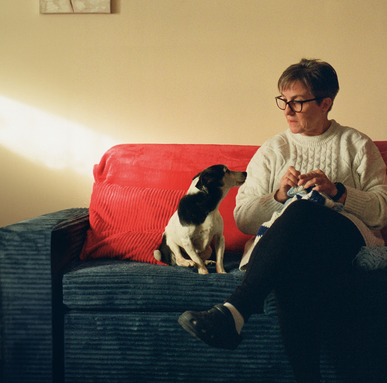 Image of living kidney donor Charlotte sitting on a red sofa. She is crocheting and looking at her dog, who is sitting on the sofa beside her and looking back at her calmly.