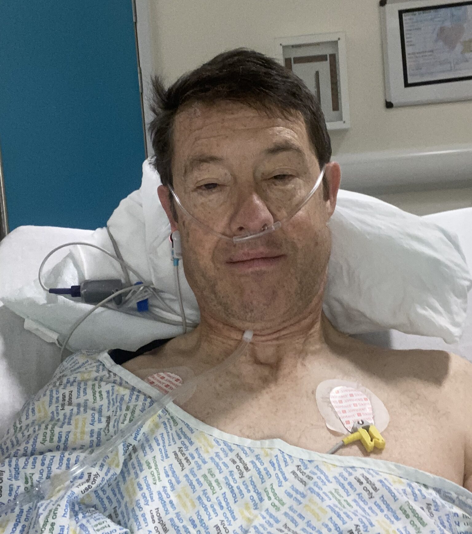 Selfie of living kidney donor Ian smiling for the camera. He is laying in a hospital bed and is smiley slightly for the camera.