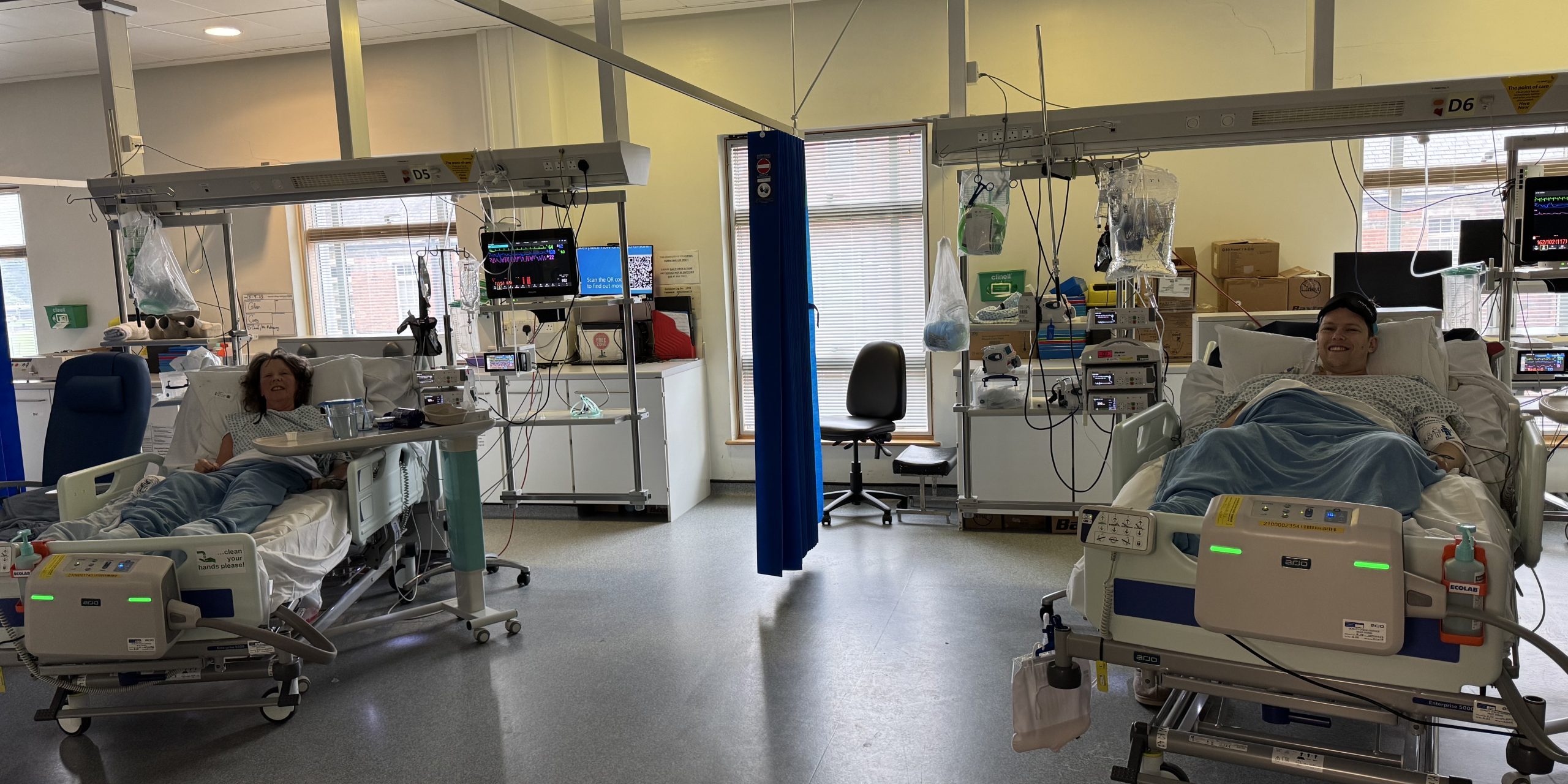 Image of living kidney recipient Luke and his kidney donor and mum. Both are laying on hospital beds beside each other, both wearing hospital gowns, and smiling for the camera.