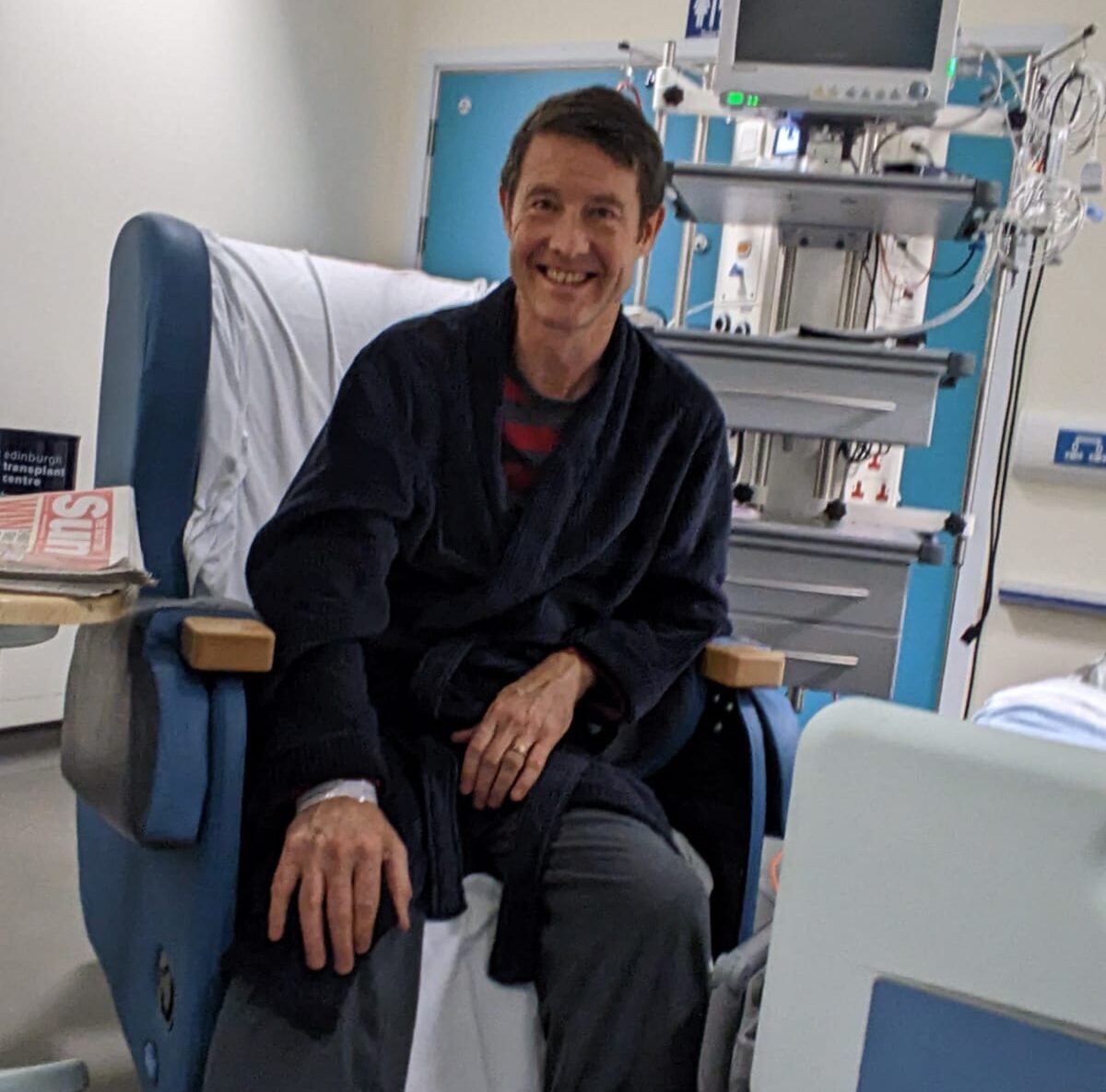 Image of living kidney donor Ian sitting on a hospital chair wearing a navy dressing gown. He is leaning on one knee and smiley widely for the camera.