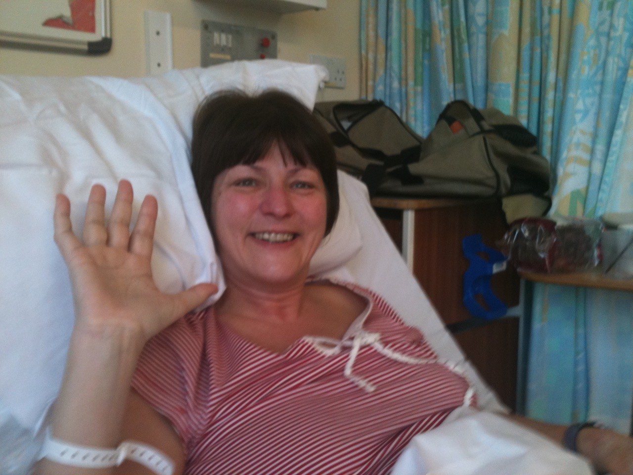 Non-directed living kidney donor Kate lying in a hospital bed wearing a red and white striped pyjama top lying in a hospital bed, smiling widely and waving to the camera. Behind her is an overnight bag on a table, in front of closed blinds.