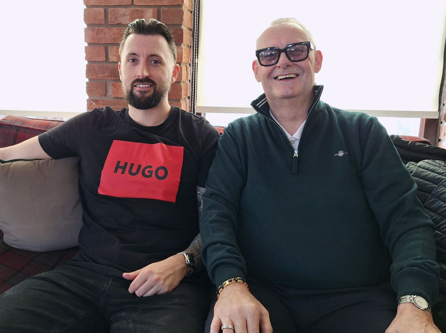 Living kidney recipient Mark alongside his son and kidney donor Aaron on a grey sofa, in front of a bright window. Mark is wearing a black jumper, while Aaron is wearing a black top with a red square on it featuring the word 'Hugo'. Both are looking at the camera and smiling