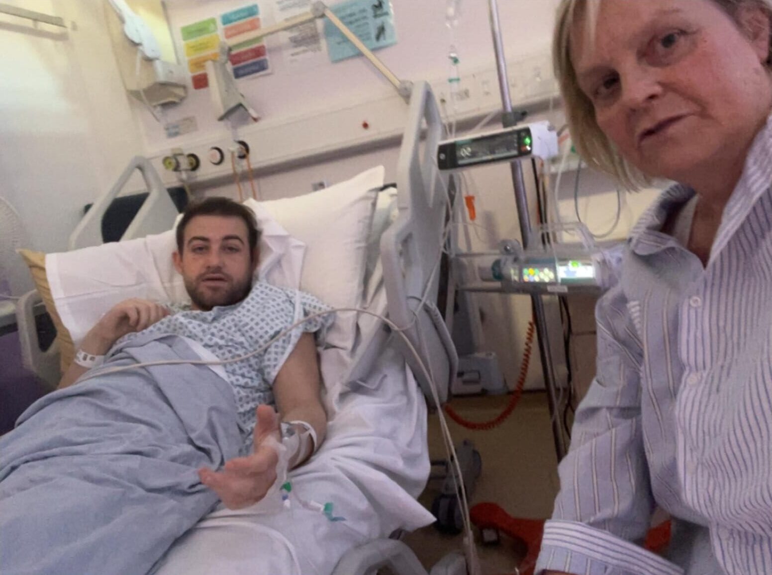 Living kidney recipient Luke lying in a hospital bed and wearing a hospital gown, looking up at the camera. Kidney donor Nicky is sitting on a chair beside his bed, wearing a blue shirt and also looking at the camera.