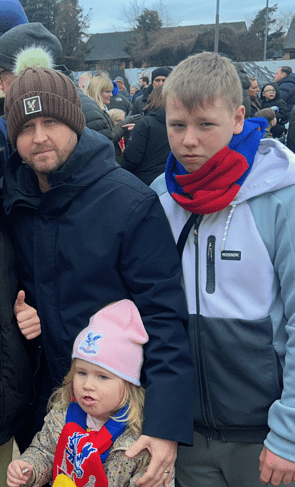 Garry standing alongside two of his children, who are wearing Crystal Palace Football Club scarves. They are all wearing thick coats on a cloudy day at the football club.