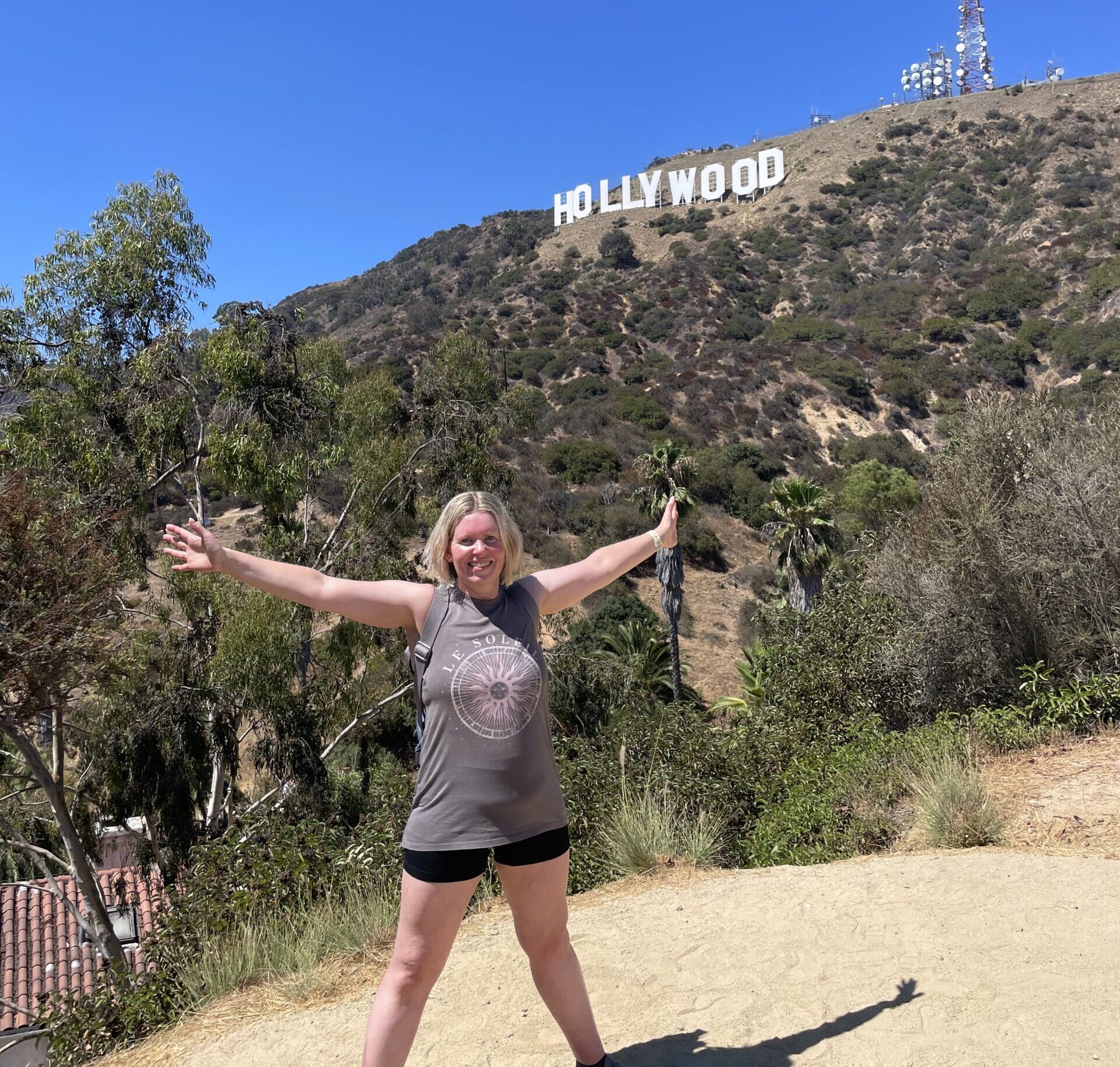 Living kidney donor Liz standing in front of the hill featuring the Hollywood sign among green trees. She is smiling with her arms outstretched wide to either side, beneath a bright blue sky.