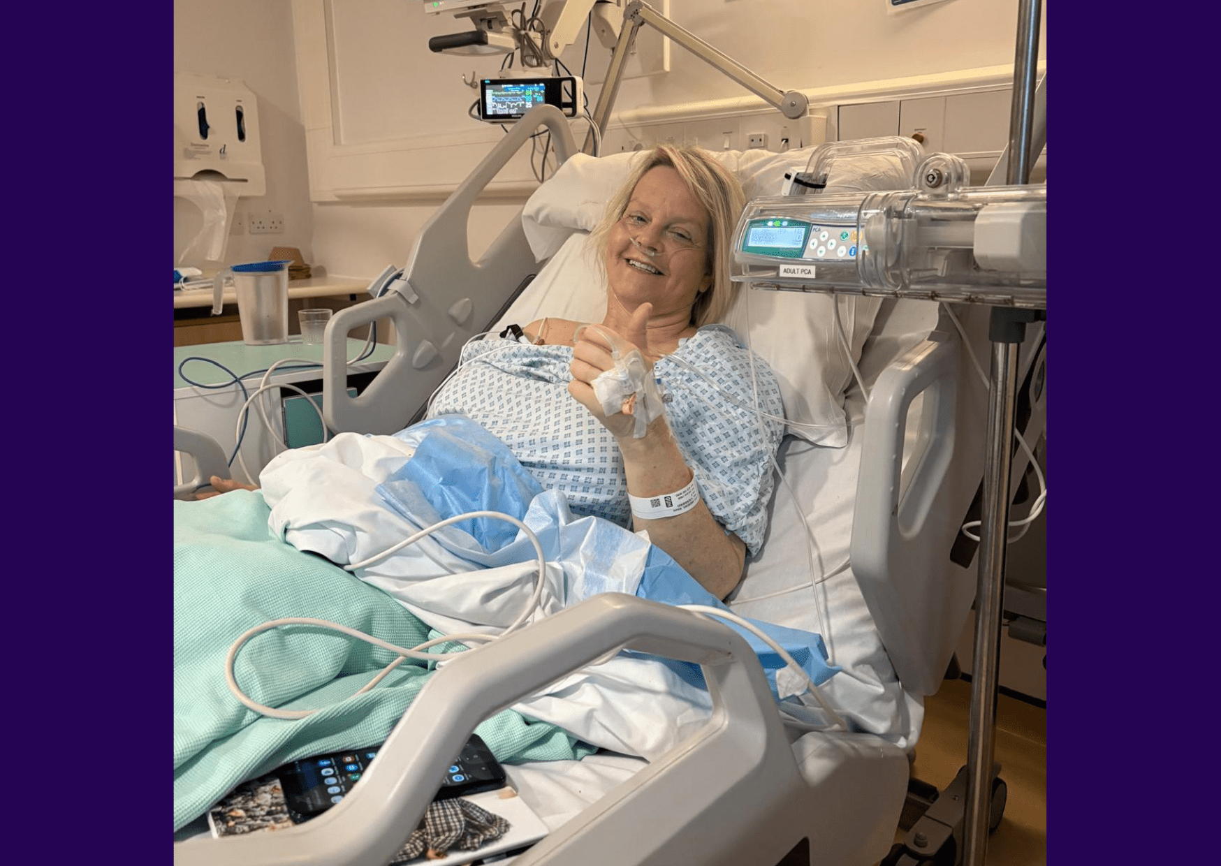 Living kidney donor Nicky lying in a hospital bed wearing a hospital gown, smiling and giving a thumbs up for the camera which is taking the photo to one side of the bed. Nicky's tablet and laptop can be seen poking out from underneath the covers beside her.