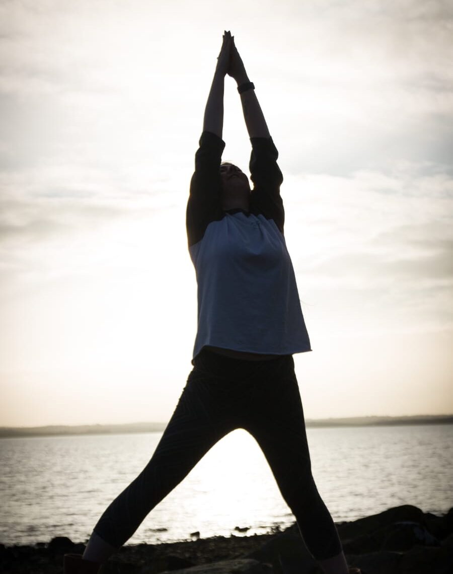 Living kidney donor Lesley standing in front of the ocean, silhouetted by the sun behind her, holding her arms high in a yoga pose.