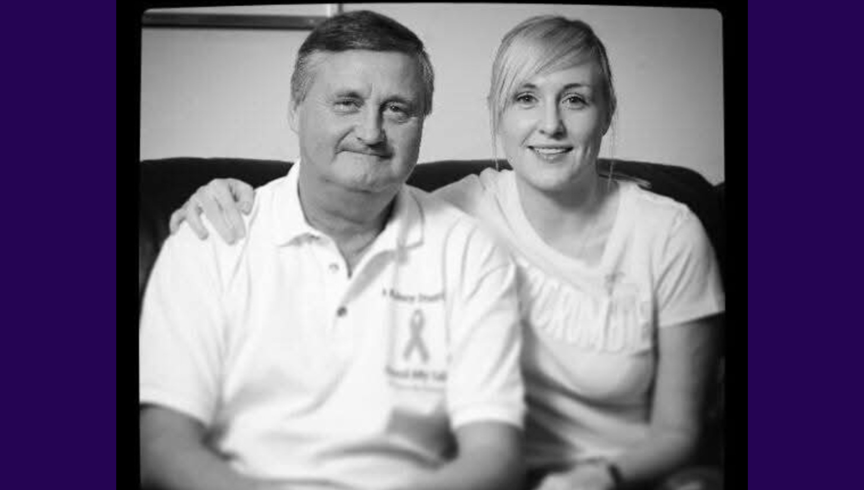 Black and white image of living kidney donor Debbie sitting alongside her father and kidney recipient. She has one hand over his shoulder and they are both smiling for the camera in front of them, both wearing white shirts. Debbie has her blonde hair tied back in a ponytail.