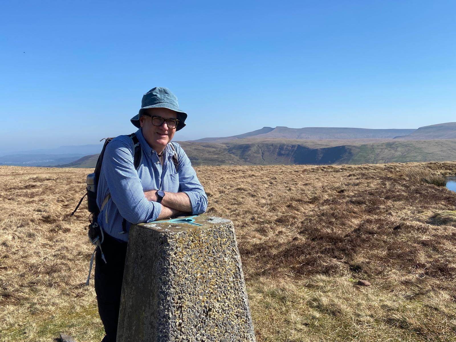 Living kidney donor Tom leaning on a walking milestone in front of a wide open grassy field, underneath a bright blue sky. He is wearing a navy bucket hat, a blue shirt, and thick-rimmed black glasses, smiling for the camera.