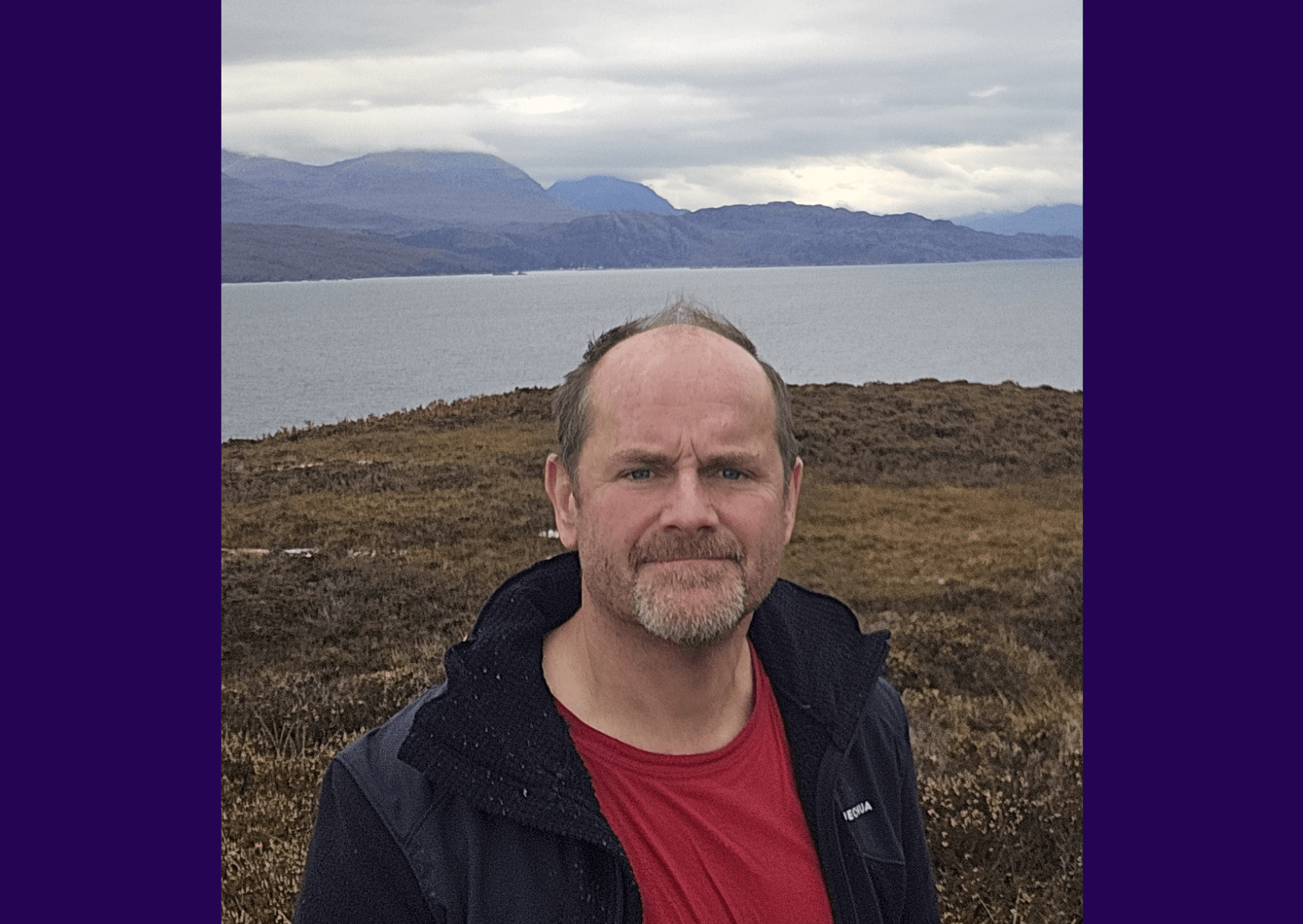 Living kidney donor Matt standing on a rocky field in front of the ocean. He is standing beneath a grey sky and is smiling for the camera. He is wearing a red top with an open navy coat.