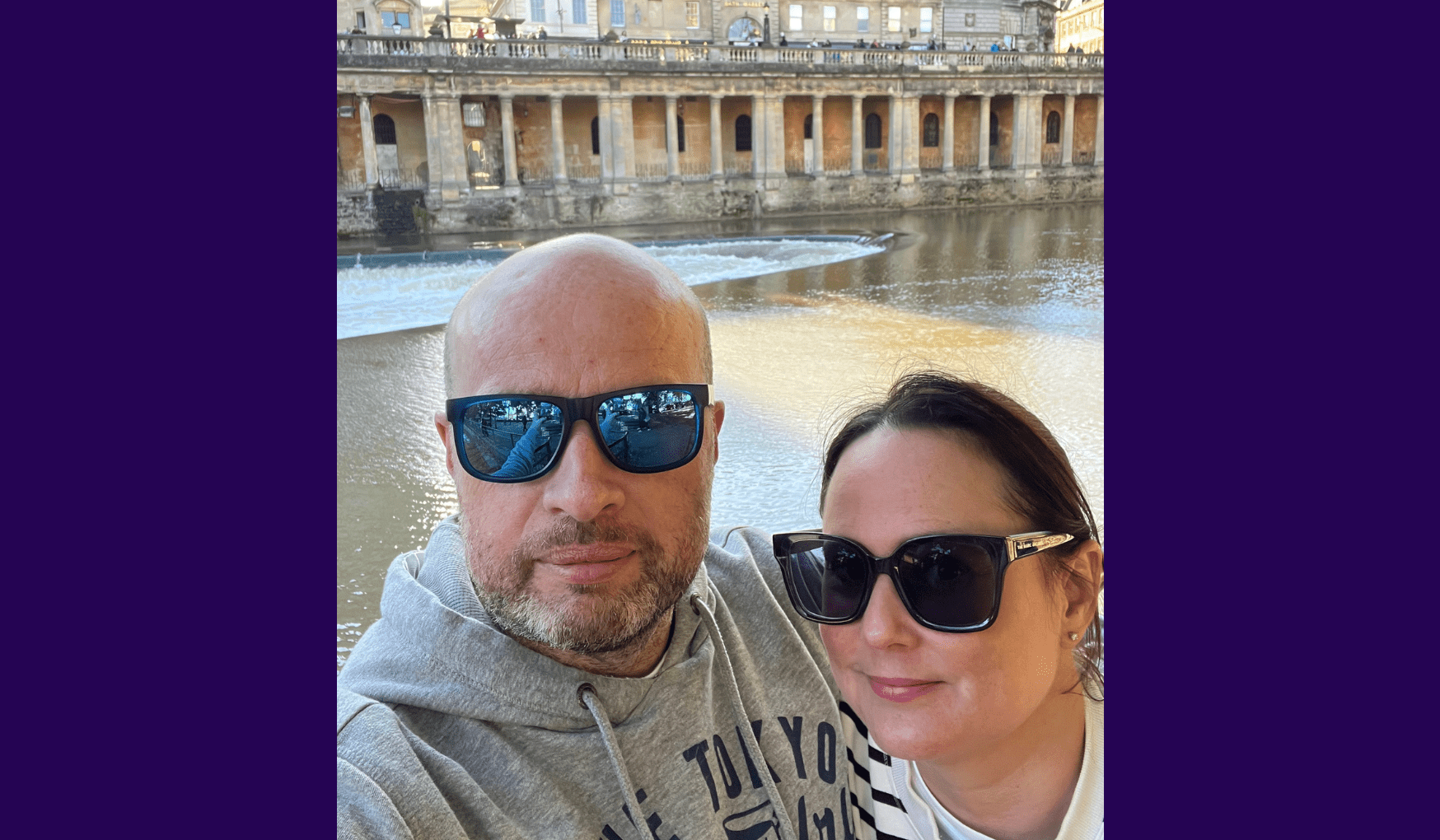 Living kidney donor Natascha standing alongside her husband Carl taking a selfie. They are both wearing dark sunglasses and standing in front of a beautiful Roman-style sandstone building on the other side of a river.