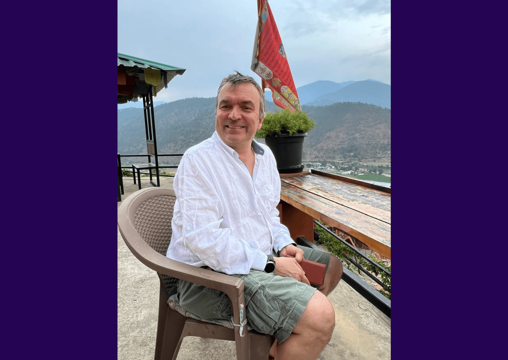 Image of living kidney donor Paul. He is wearing a loose white shirt and light green shorts, sitting on a chair looking over a valley, his phone in his hands, looking towards the camera and smiling.