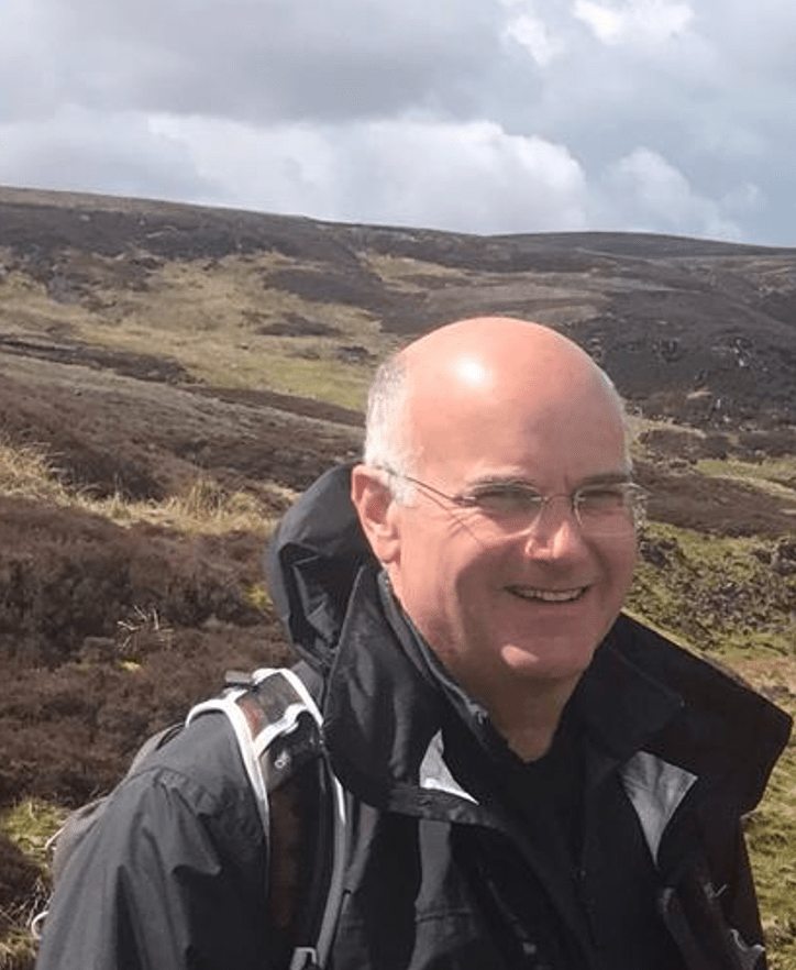 Living kidney donor Tom on a walking holiday, standing in front of a wide open plain beneath an overcast sky. He is smiling for the camera and wearing a black raincoat.