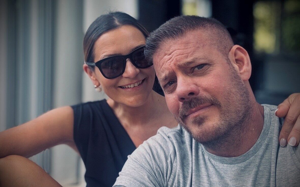 Image of living kidney donor Lisa standing beside her husband and kidney recipient, in front of a blurred background. She is wearing sunglasses and a black top, smiling widely, while her husband is wearing a grey top and leaning his head towards her.