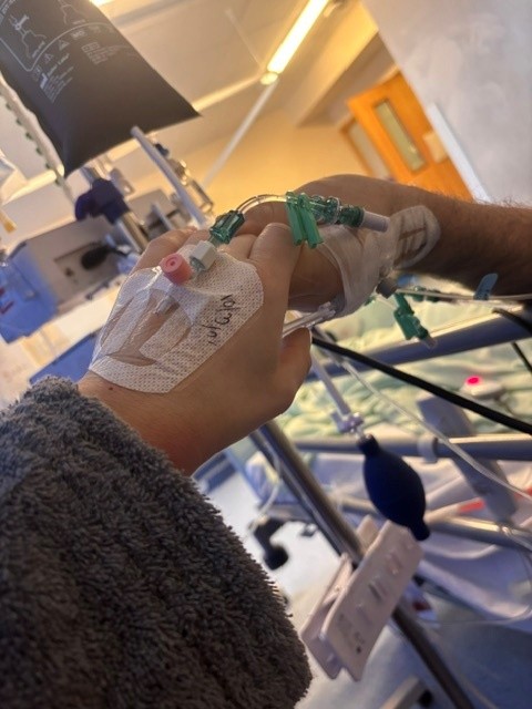 A close up of two hands holding each other in a hospital. A bag of blood is visible just behind them, and both hands have a cannula attached to them.