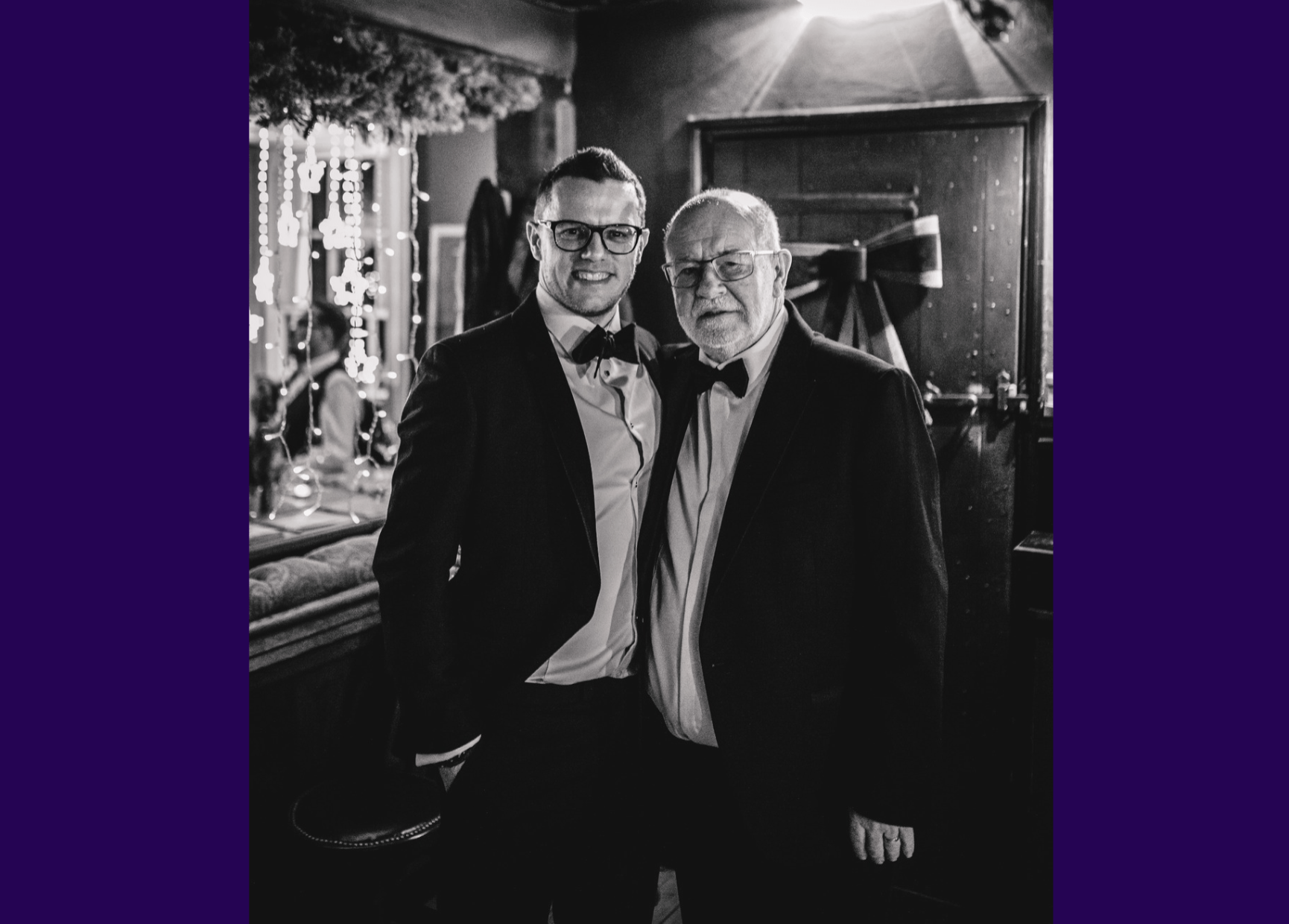 Black and white image of living kidney donor Luke alongside his dad and kidney recipient. They are both wearing tuxedos with bow ties in front of a bar and are smiling for the camera.