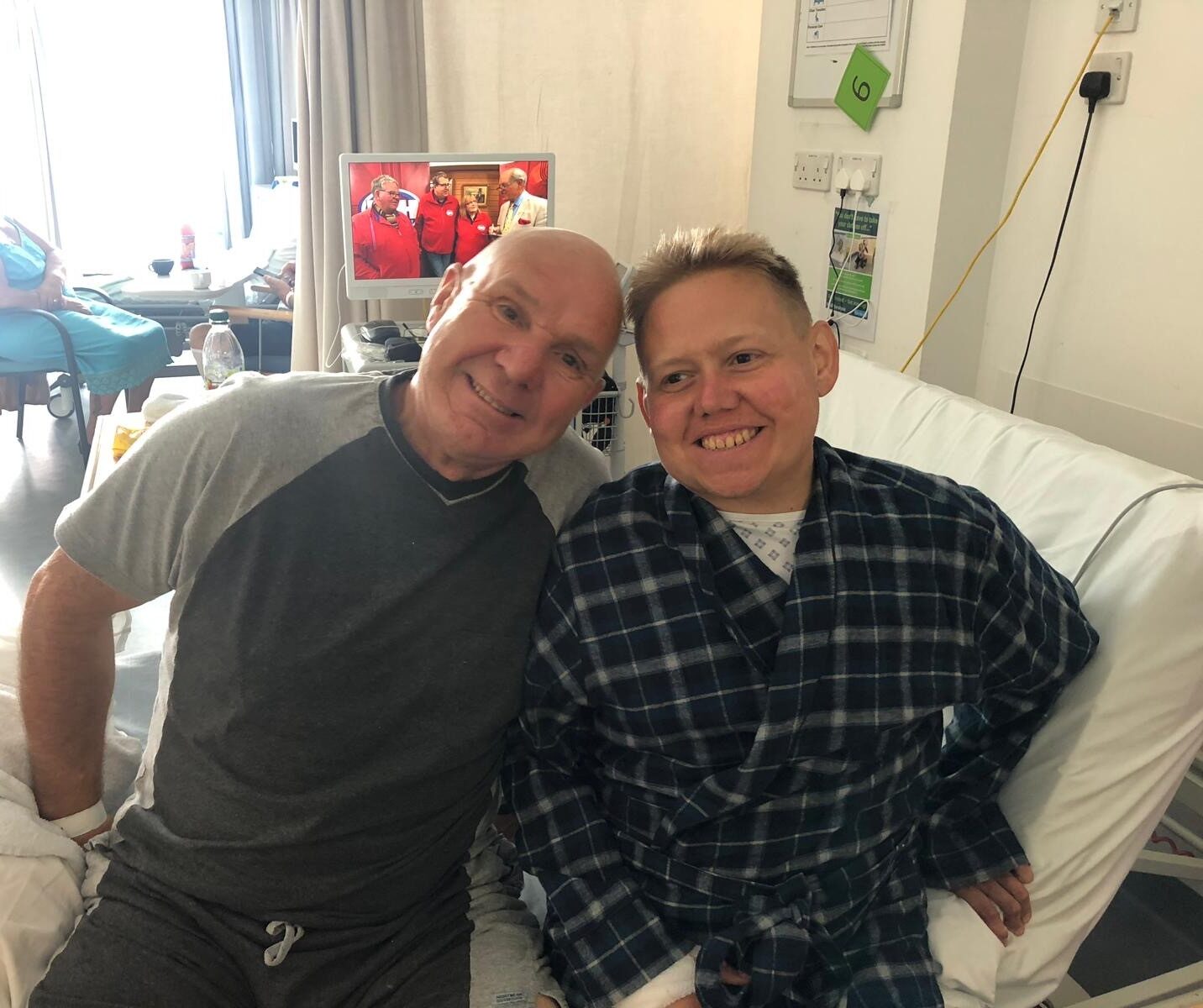 Image of living kidney donor standing alongside his stepson and recipient. Tony has his arm around his stepson. They are sitting on a hospital bed and smiling widely at the camera, heads leaned together next to each other.