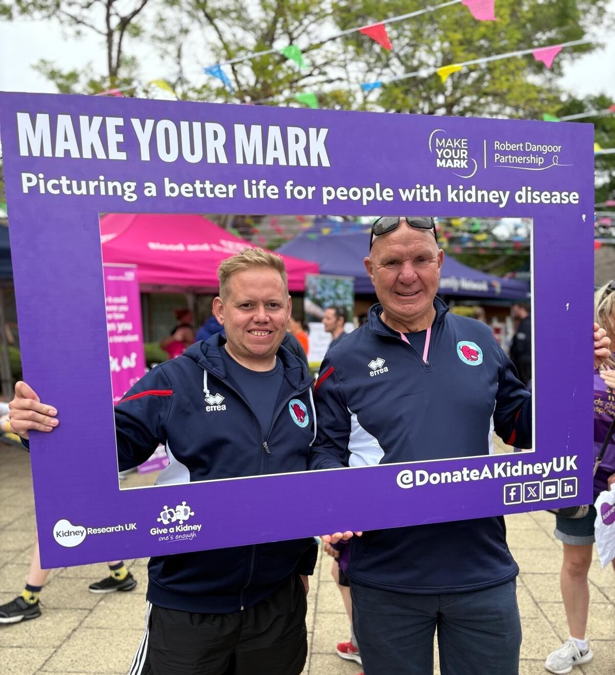 Living kidney donor Tony and his stepson and recipient standing inside a selfie frame branded with the Make Your Mark campaign from the Robert Dangoor Partnership for Living Kidney Donation. They are both wearing running clothes and are smiling for the camera.