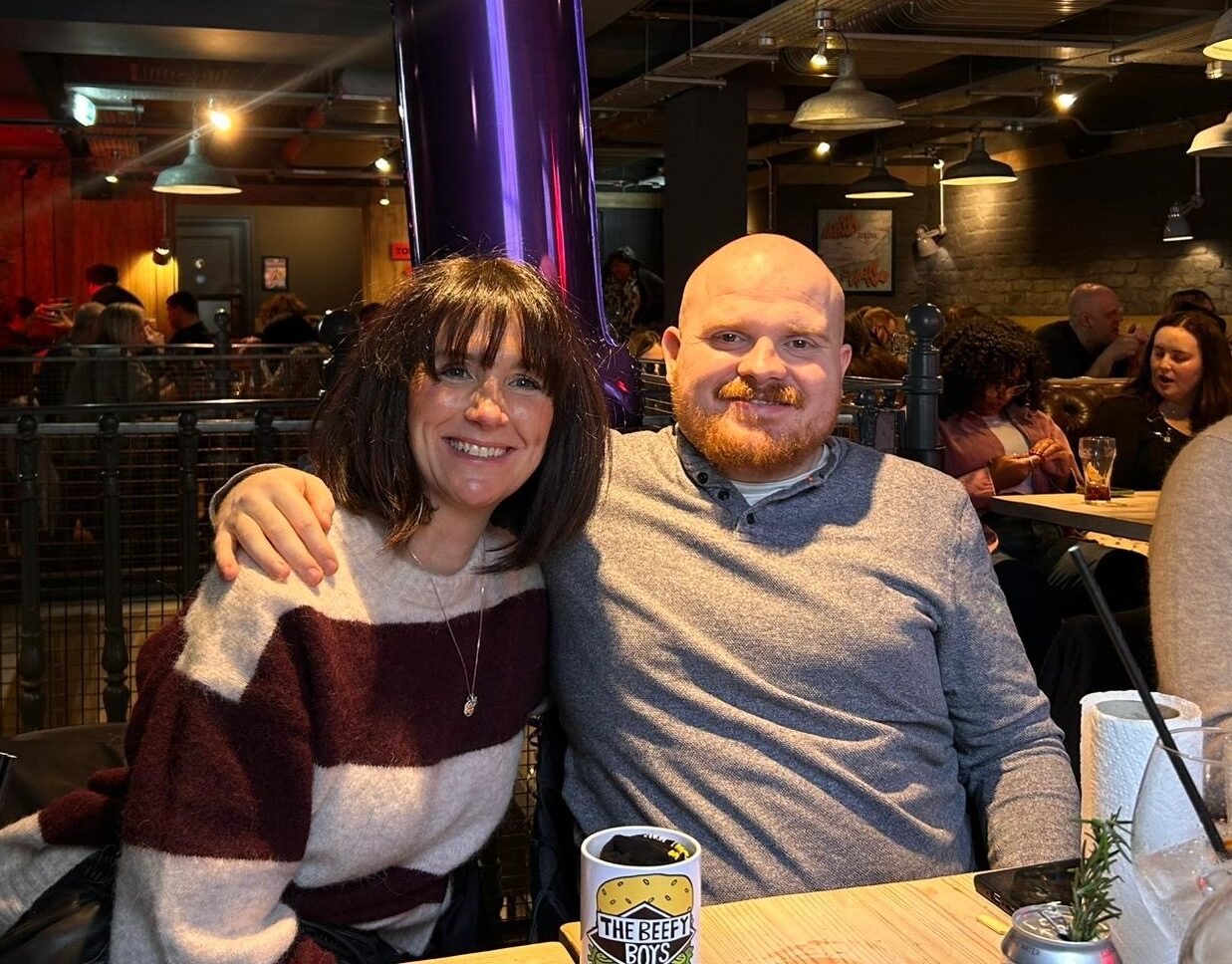 Image of living kidney donor Jo and her nephew Louis, sitting in a restaurant next to each other. Louis has his arm over Jo's shoulder, hugging her towards him. She is wearing a grey and black striped jumper, while Louis is wearing a grey top. They are both smiling widely.