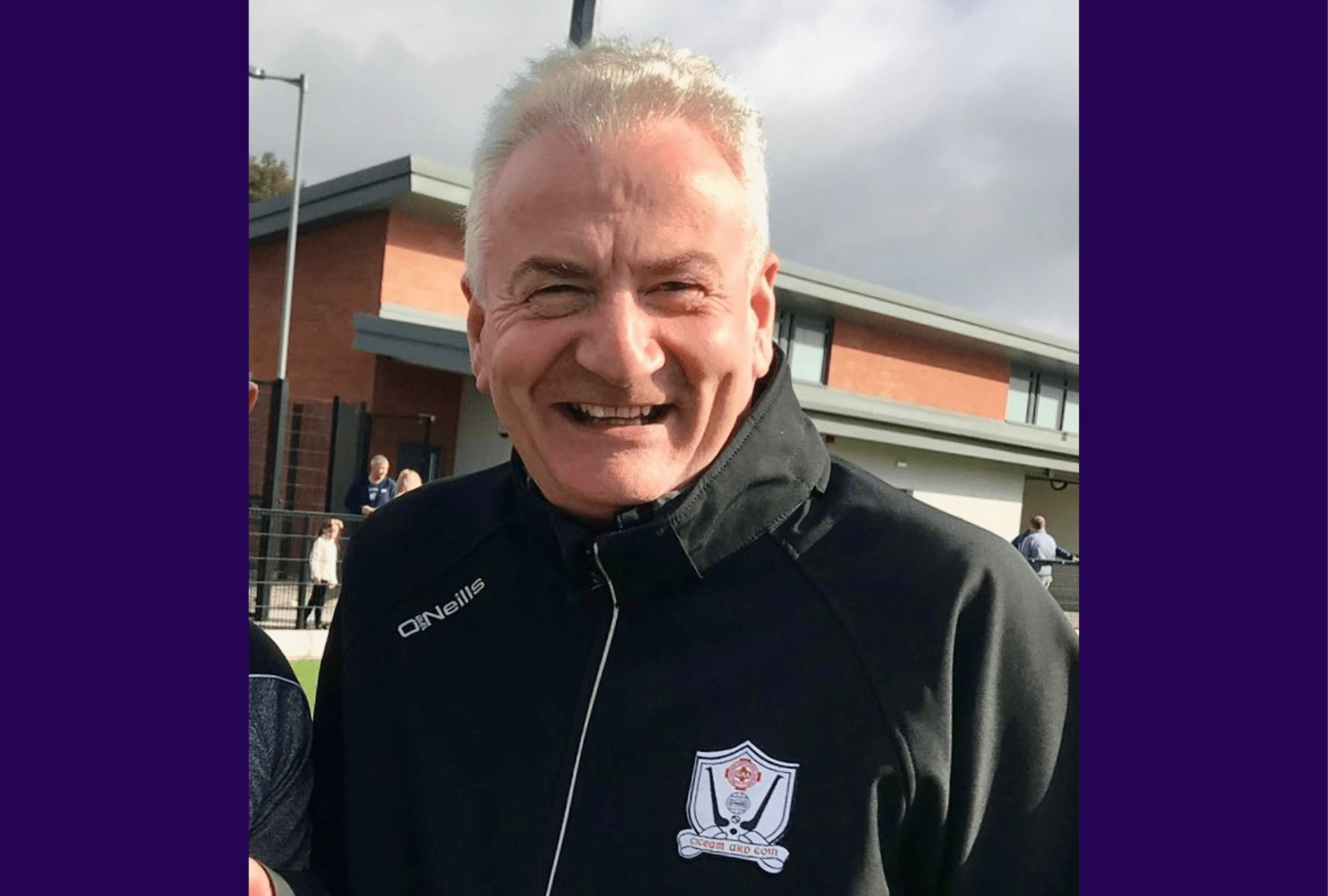Image of living kidney donor widely smiling for the camera. A grey cloudy sky is visible behind him. He is wearing a black Gaelic football fleece.