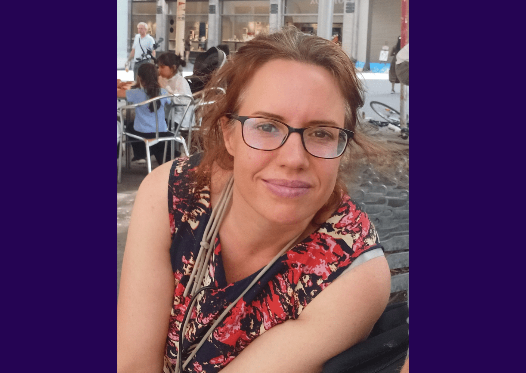 Image of living kidney donor Wilma sitting at a table outside, leaning towards the camera and smiling. She is wearing a black dress with red and cream splashes of colour on it, as well as black rimmed glasses. A number of other outdoor restaurant tables can be seen behind her.